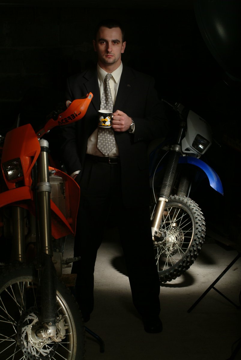 Michał Bieliński w garniturze z kubkiem kawy między dwoma motocyklami enduro